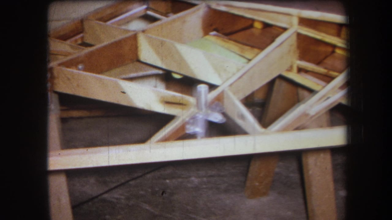 1964 8mm frame from Yucaipa, California, showing pale wooden framing, diagonal braces, and metal fasteners on a workshop floor.