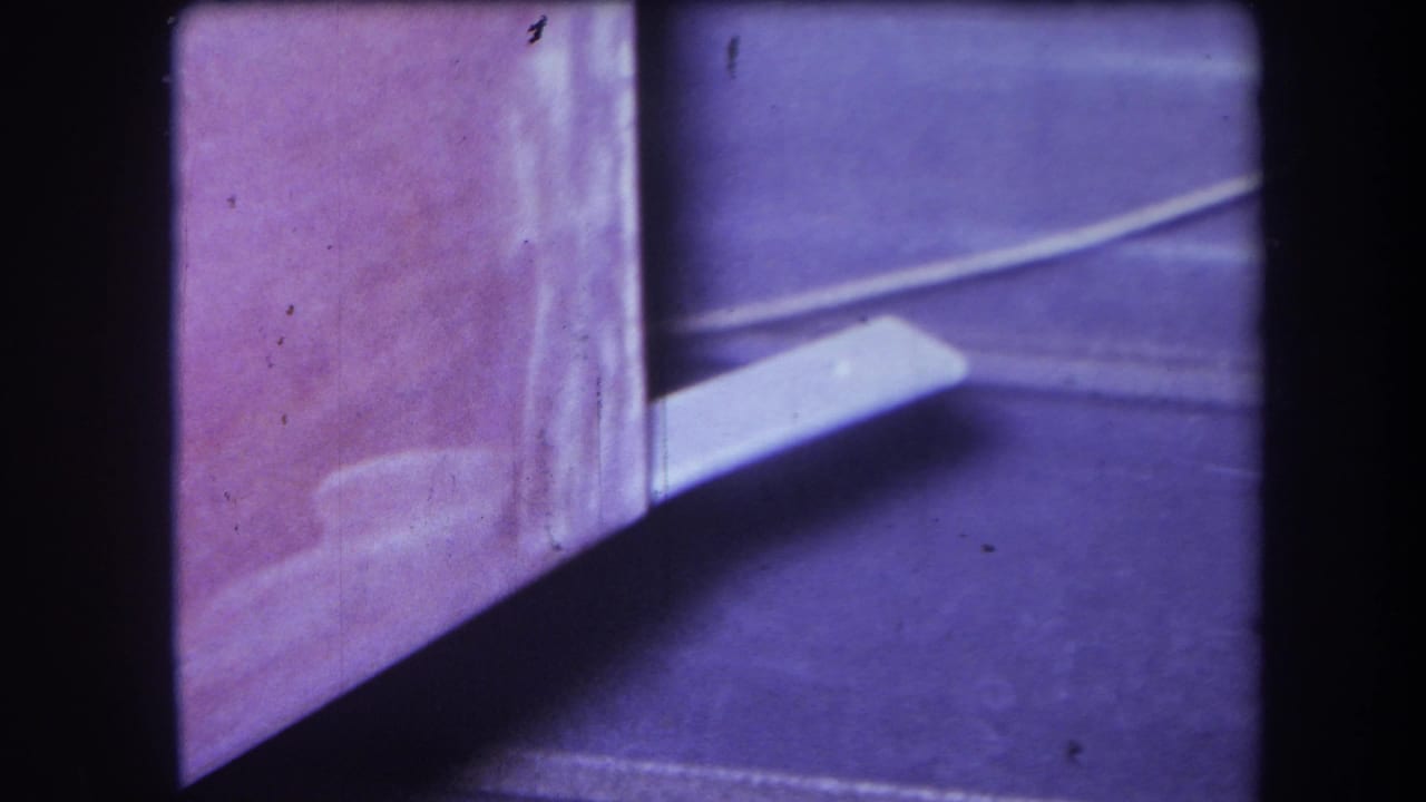 1964 Yucaipa 8mm frame showing a white rectangular object extending from a pink-purple corner surface with shadowed floor area.