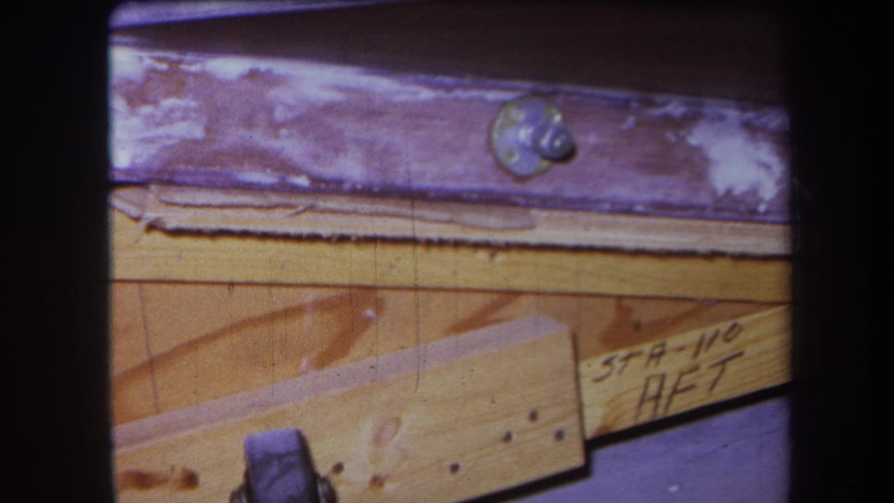 Close 1964 8mm frame of workshop woodwork, showing layered planks, visible grain, a round fastener, nail points, and handwritten construction notation.