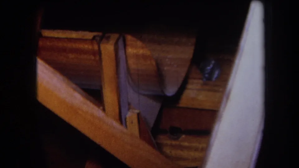 1964 8mm close-up of a wooden framework or craft assembly, with an angled rail, darker support piece, fastener, and pale curved plastic or fiberglass-looking panel.