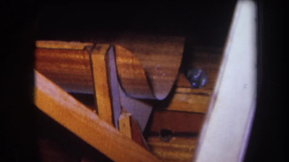 Close 1964 8mm view of workshop construction details, including varnished wood framing, a dark curved support, small fastener, and pale smooth panel at the side.