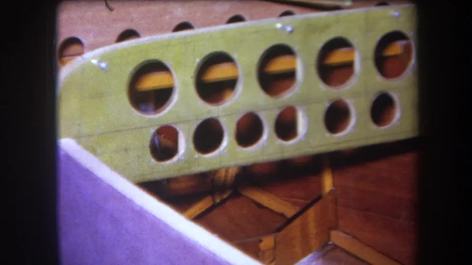 1960s home-movie frame of a green painted wooden panel seen at an angle, with round holes and reddish wood construction below.
