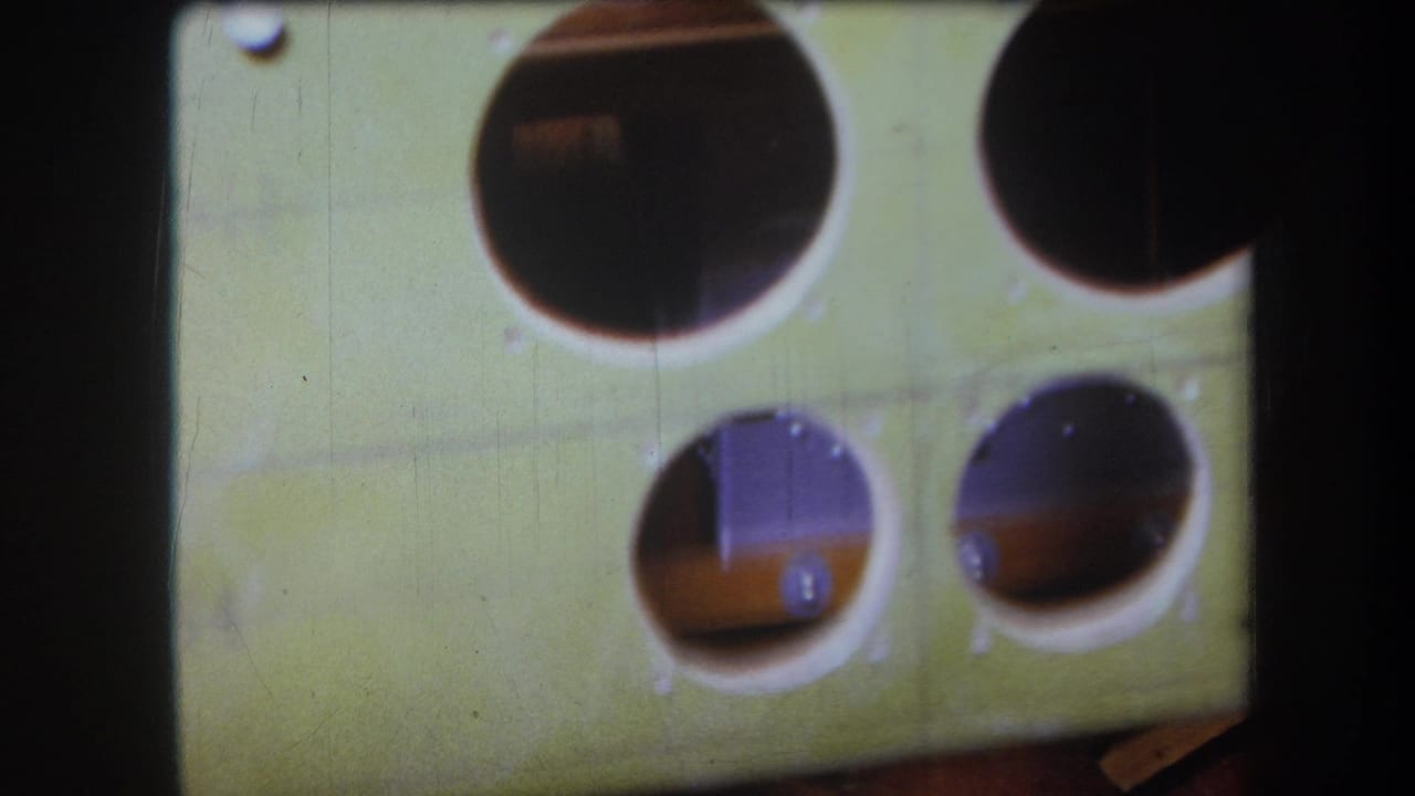 Close 8mm view of the green wooden instrument-like panel, with large round openings, smaller holes, screws, and shadowed interior wood.