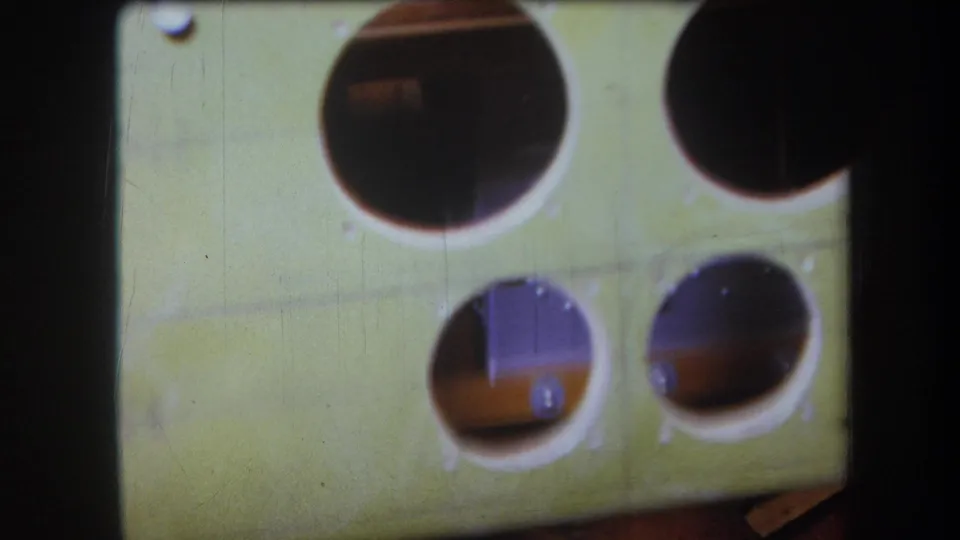 Close 8mm view of the green wooden instrument-like panel, with large round openings, smaller holes, screws, and shadowed interior wood.