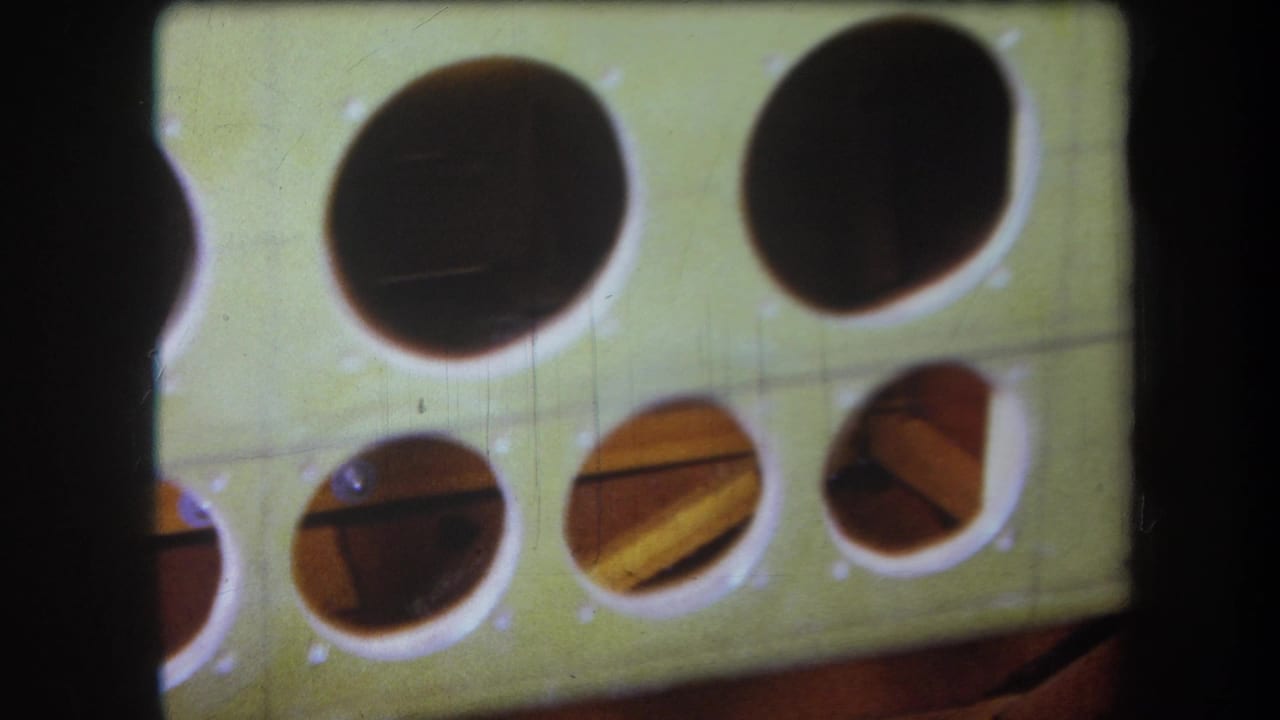 1964 Volmer Yucaipa home-movie frame showing a pale green board or template with round cut openings and visible film grain.