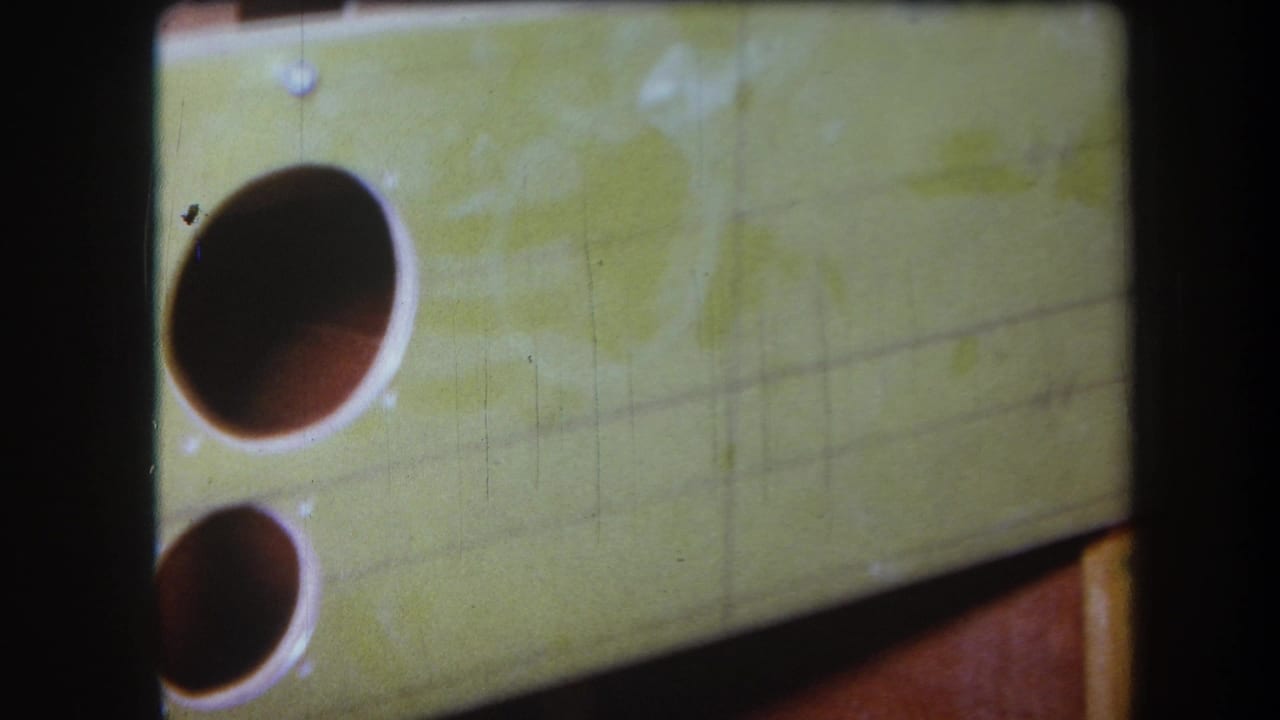 1964 8mm frame showing the same yellow wall or panel with two dark round openings, tilted composition, and visible film scratches.