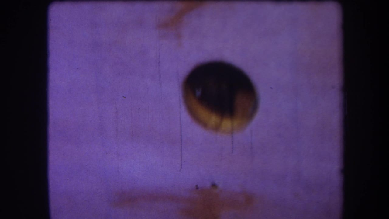 1964 8mm home-movie frame from the supplied Yucaipa reel, showing a dark circular aperture against purple faded film with scratches and dust.