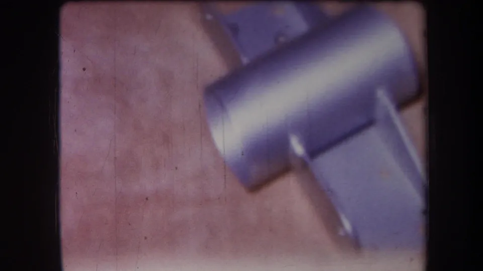 1964 8mm workshop close-up from Yucaipa showing a small blue-gray metal cylinder and attached flat pieces on the bench.