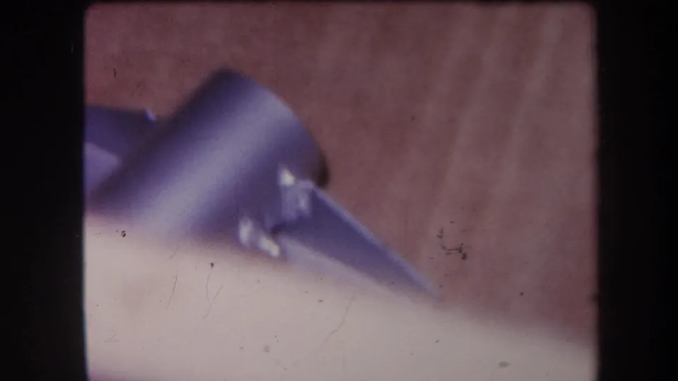Close 1964 8mm view of a purple-blue metal fixture or bracket on wood, with a pale foreground edge and film scratches.