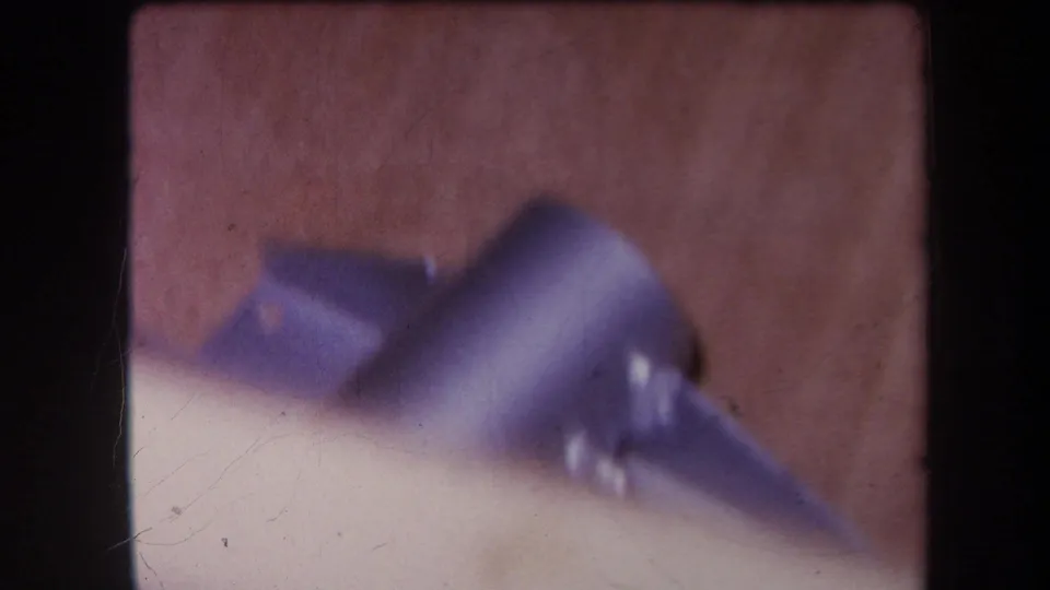 Final 1964 8mm frame showing a purple-blue metal fixture on a wooden surface, with scratches, grain, and dark film borders.