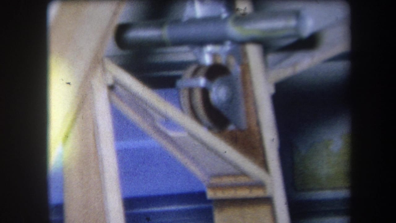1964 8mm frame from Yucaipa-area material showing metal gears or pulleys on an angled wooden mechanical structure.