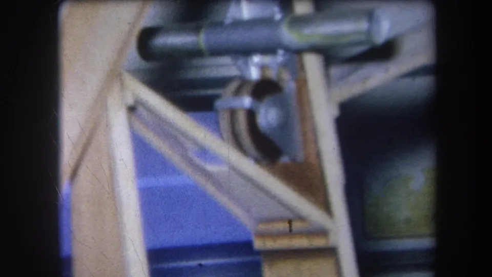 1964 8mm frame showing a close, slightly faded view of metal gears or pulleys attached to a light wooden structure.