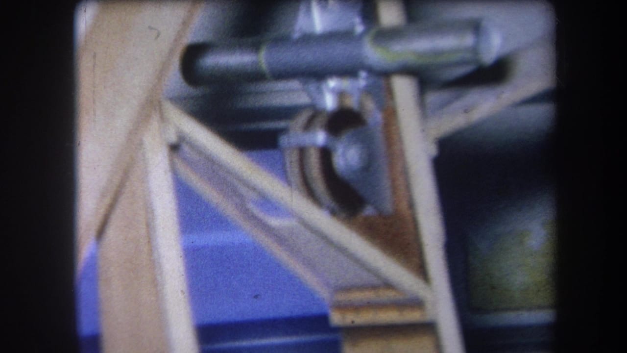 Close 8mm view of a wooden-and-metal mechanism with angled supports, rod hardware, and a dark pulley-like wheel.