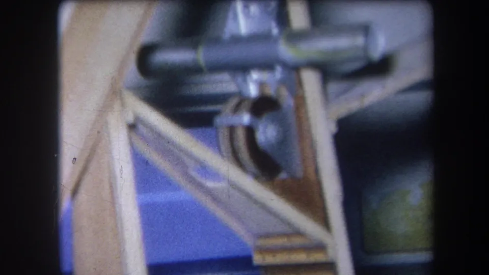 Soft 1964 8mm frame showing the same wooden machinery support, metal joint, and blue backing in close view.