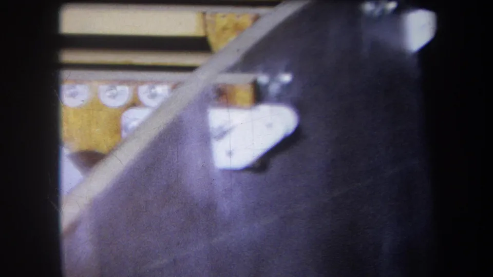 Close 1964 8mm view of industrial machine components, including white bracket shapes, yellow-painted parts, and dark metal housing.