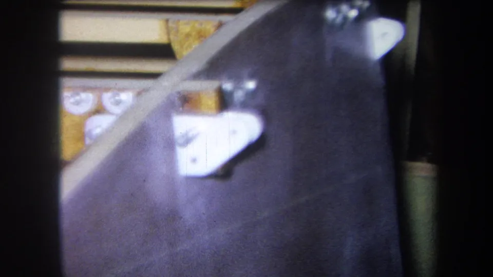 1964 Yucaipa-related 8mm frame showing a white metal bracket, yellow machinery parts, dark surface, and visible film softness.