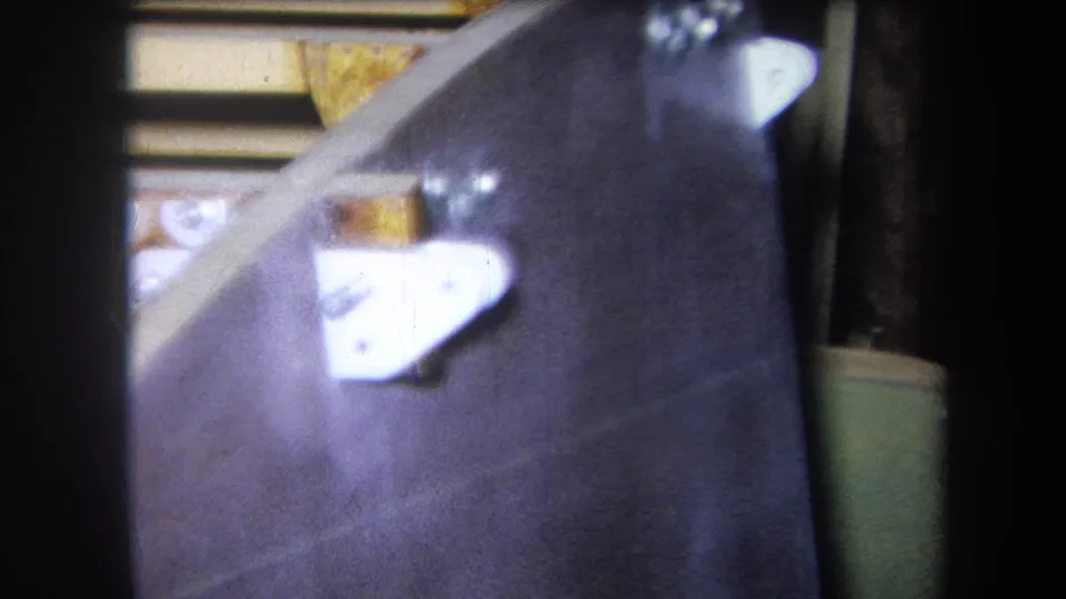 Close 8mm frame from the 1964 metadata showing dark industrial machinery, a white bracket, yellow component, and edge artifacts from film scanning.