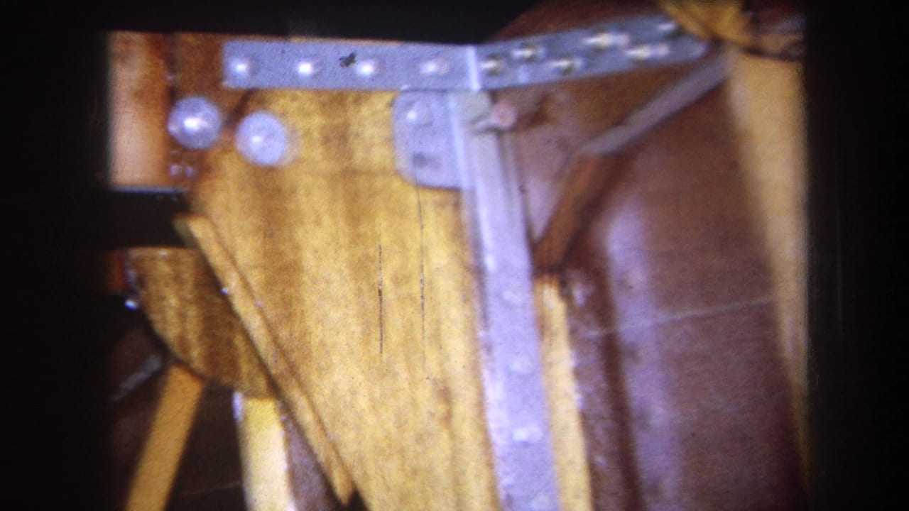 1964 Yucaipa home-movie frame showing the underside of a wooden table with metal hardware, pale support pieces, and a brown finished surface.