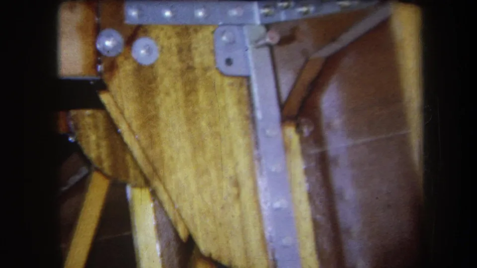 1964 Yucaipa home-movie view under a table, with angled supports, metal bracket hardware, bolts, and varnished brown material visible.