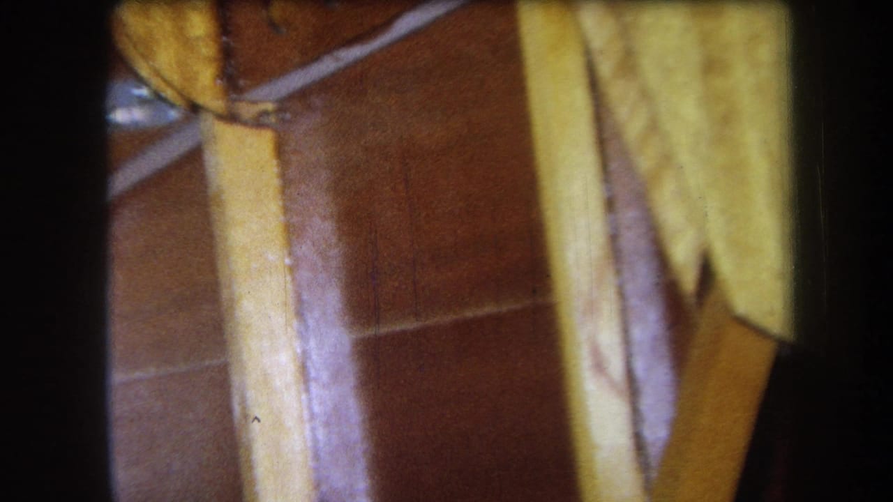 1964 Yucaipa home-movie frame showing a close underside view of varnished wooden furniture with rails and warm brown tile in the background.