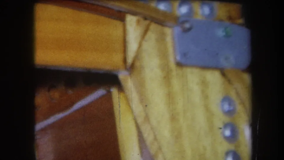 1964 Yucaipa home-movie close-up of a wooden furniture underside, with a gray metal plate, screw heads, diagonal supports, and tile behind it.