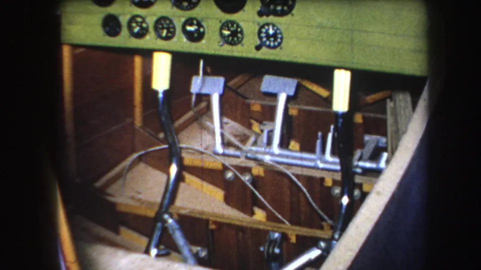 1964 8mm cockpit view with a green dashboard, clustered round instruments, metal pedals, control rods, wiring, and wood cabin panels.