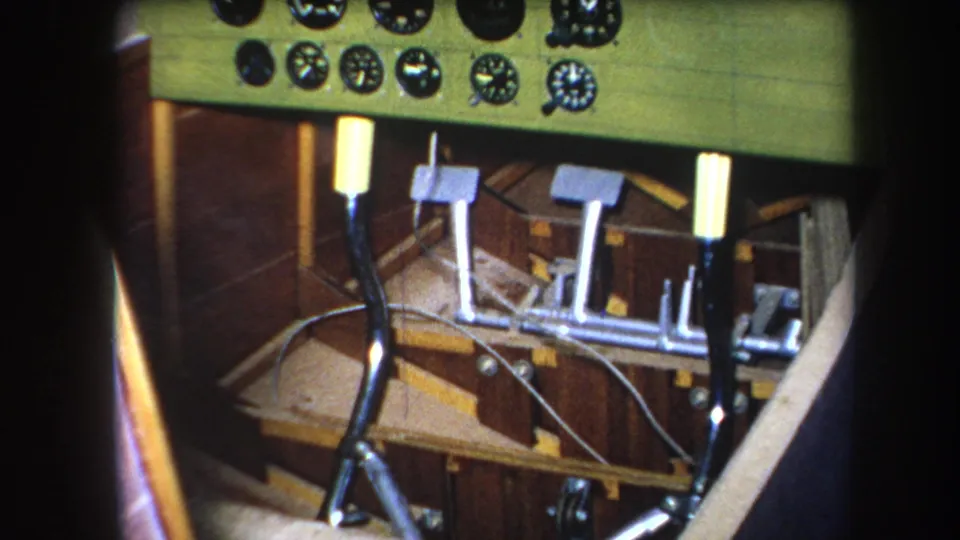 Close 1964 home-movie frame of aircraft controls with analog gauges, upright levers, pedals, loose cables, and warm-toned wood flooring.
