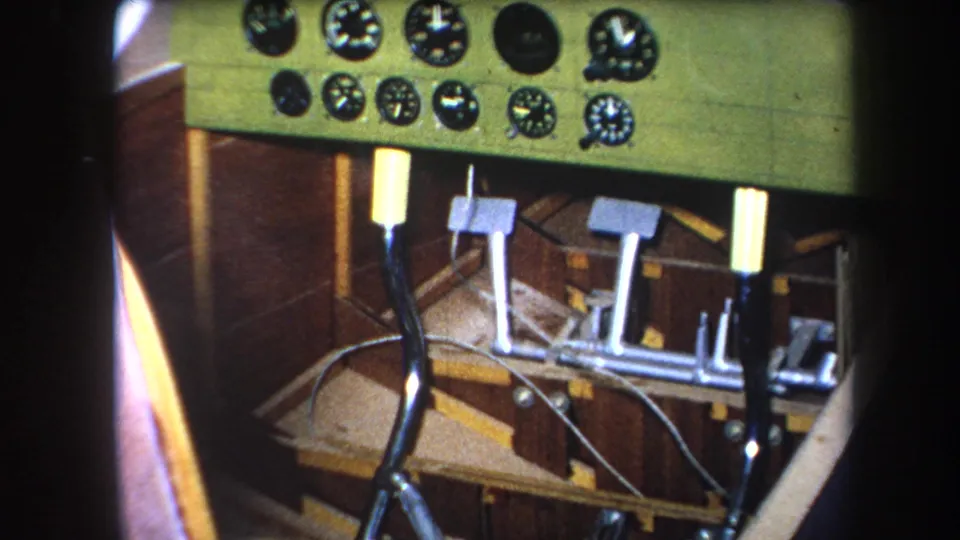 Final 1964 8mm frame showing the small aircraft cockpit interior, including green gauge panel, pedal assembly, exposed cables, and wooden cabin structure.