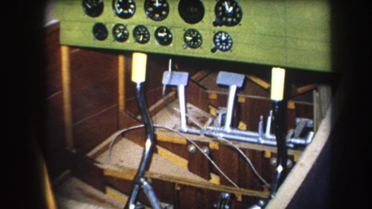 Close-up 1964 home-movie frame of an aircraft cockpit in Yucaipa, California, with a green gauge panel, metal pedals, joystick, wiring, and wood structure.