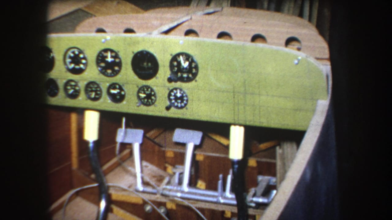 1964 8mm frame from Yucaipa area footage showing a green homemade aircraft cockpit panel with gauges, pedals, wiring, and wood framing.