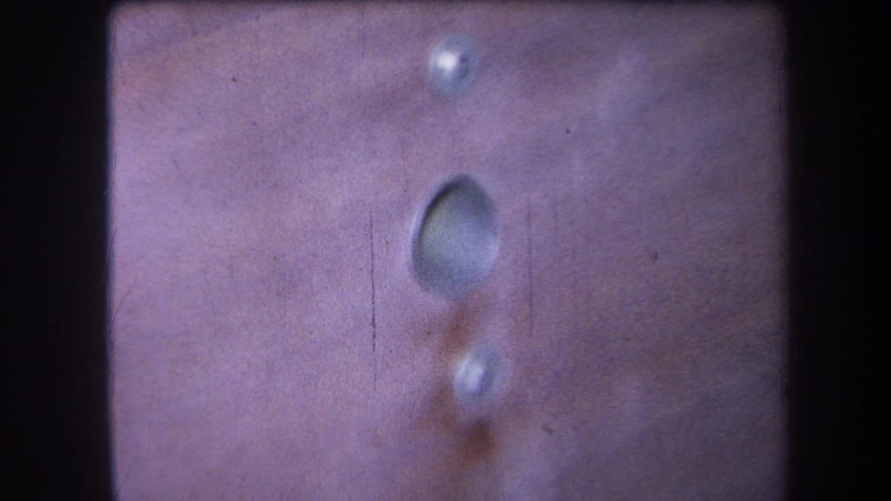 Close 1964 8mm view of a pinkish riveted metal surface, with a recessed oval mark and visible film grain.