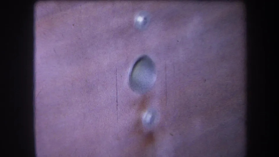 Close 1964 8mm view of a pinkish riveted metal surface, with a recessed oval mark and visible film grain.