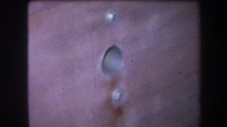 1964 8mm close-up showing a rivet, recessed oval mark, and reddish stain on a metal surface in handheld framing.