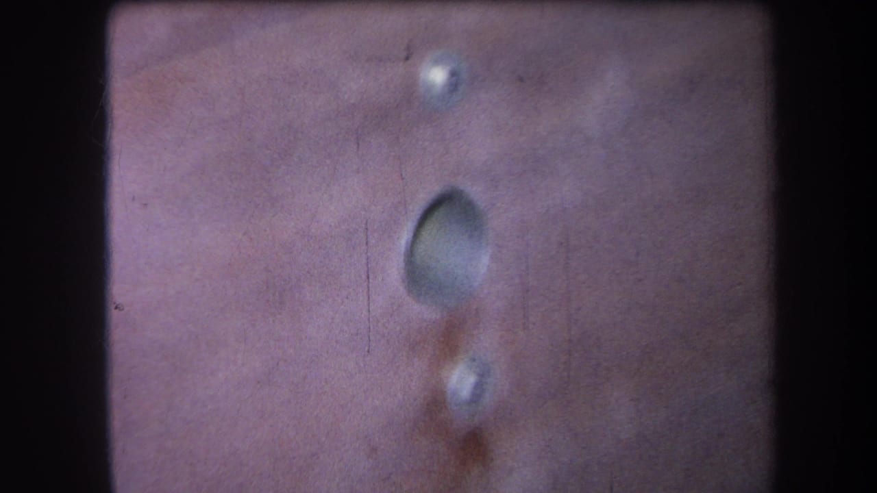 1964 8mm close-up from Volmer Yucaipa showing a wooden panel with a central round hole, small fastener holes, grain, and film-edge shadow.