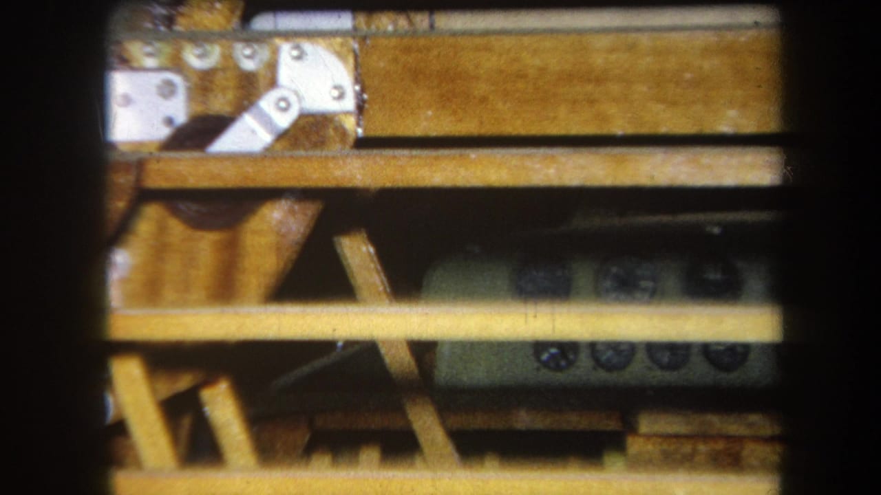 1964 Yucaipa home-movie frame showing a green-gray control device partly obscured by wooden slats and table hardware.