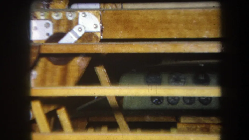 1964 Yucaipa home-movie frame showing a green-gray control device partly obscured by wooden slats and table hardware.