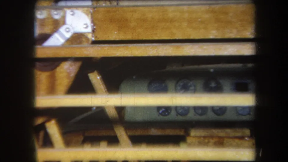 Final 1964 Yucaipa still of a remote-like control device under a wooden table, framed by slats, brackets, and dark film edges.