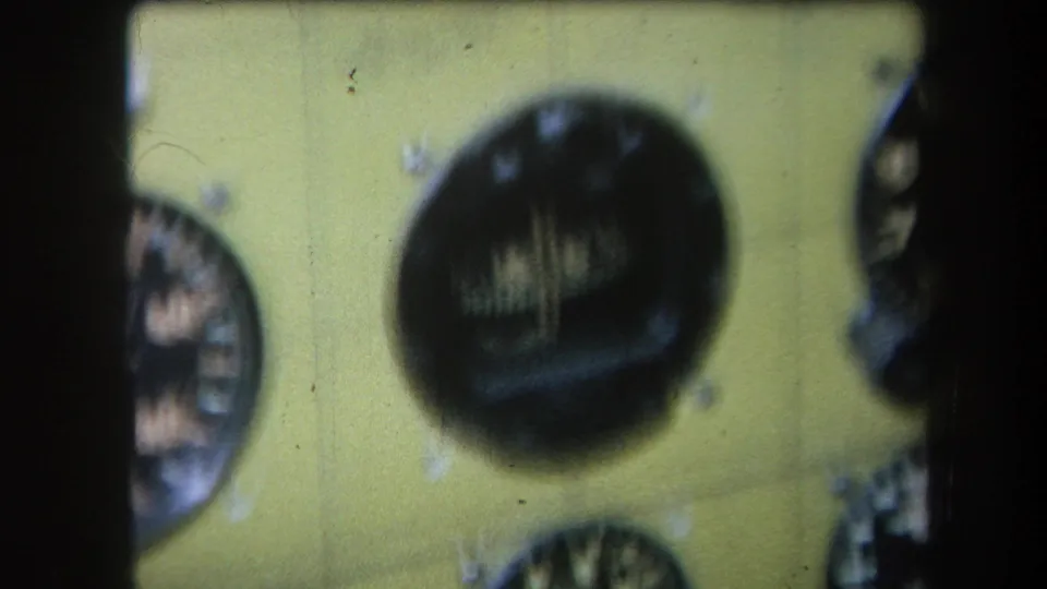 Close 1964 8mm view of a yellow instrument panel, with a large dark analogue dial and partial circular gauges near the frame edges.