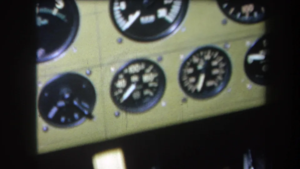 1964 home-movie frame showing lower cockpit gauges on a green aircraft panel, with soft focus and dark shadowed edges.