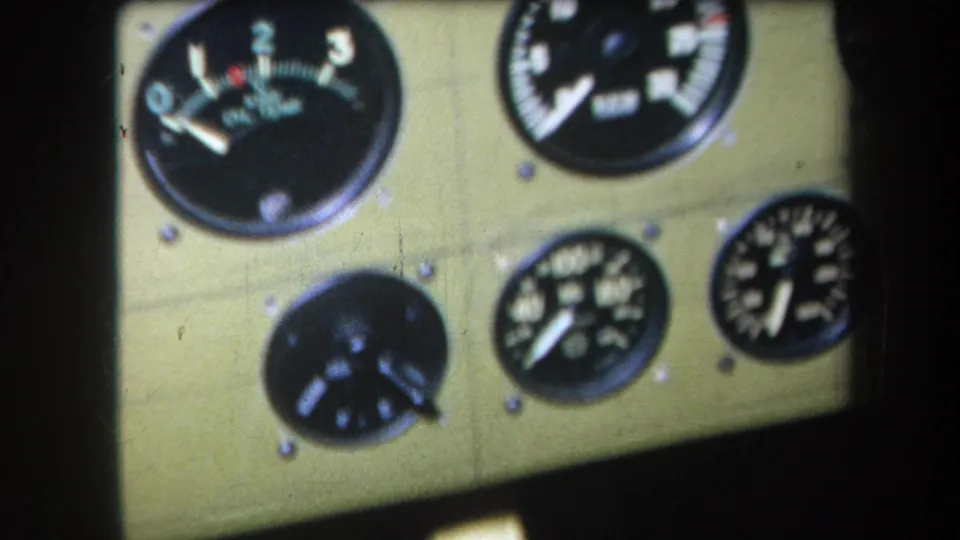 1964 home-movie frame showing a green cockpit dashboard with multiple analog gauges, including a dial marked with numbers and a red arc.