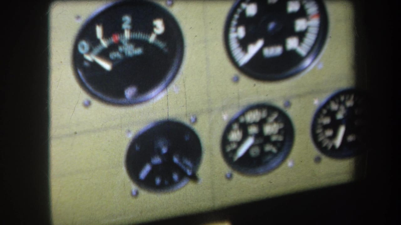 1964 8mm close-up of black-faced analog dials mounted on a khaki panel, with soft focus and dark edge shadow.