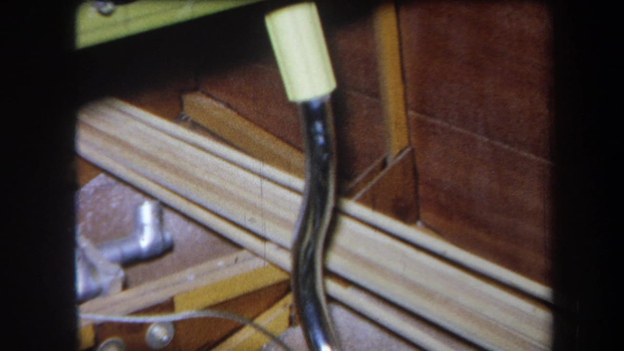 1964 Yucaipa home-movie frame showing a close view of a wooden board with metal rods, hardware, black cable, and yellow connector.