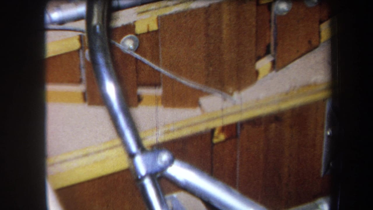 1964 Yucaipa home-movie frame showing silver metal tubing, yellow-painted rails, and brown wooden panels in close-up.