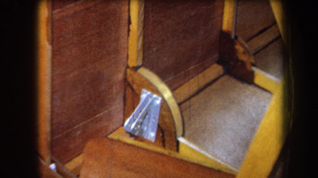 1964 8mm frame from Yucaipa showing brown wood panels, yellow framing, a gray floor area, and a silver bracket in a small constructed interior.