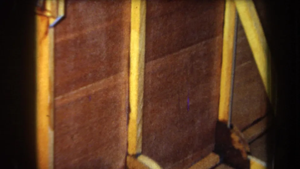 1964 Yucaipa home-movie frame showing brown boards, a yellow vertical strip, and a dark curved film edge beside part of a small structure.