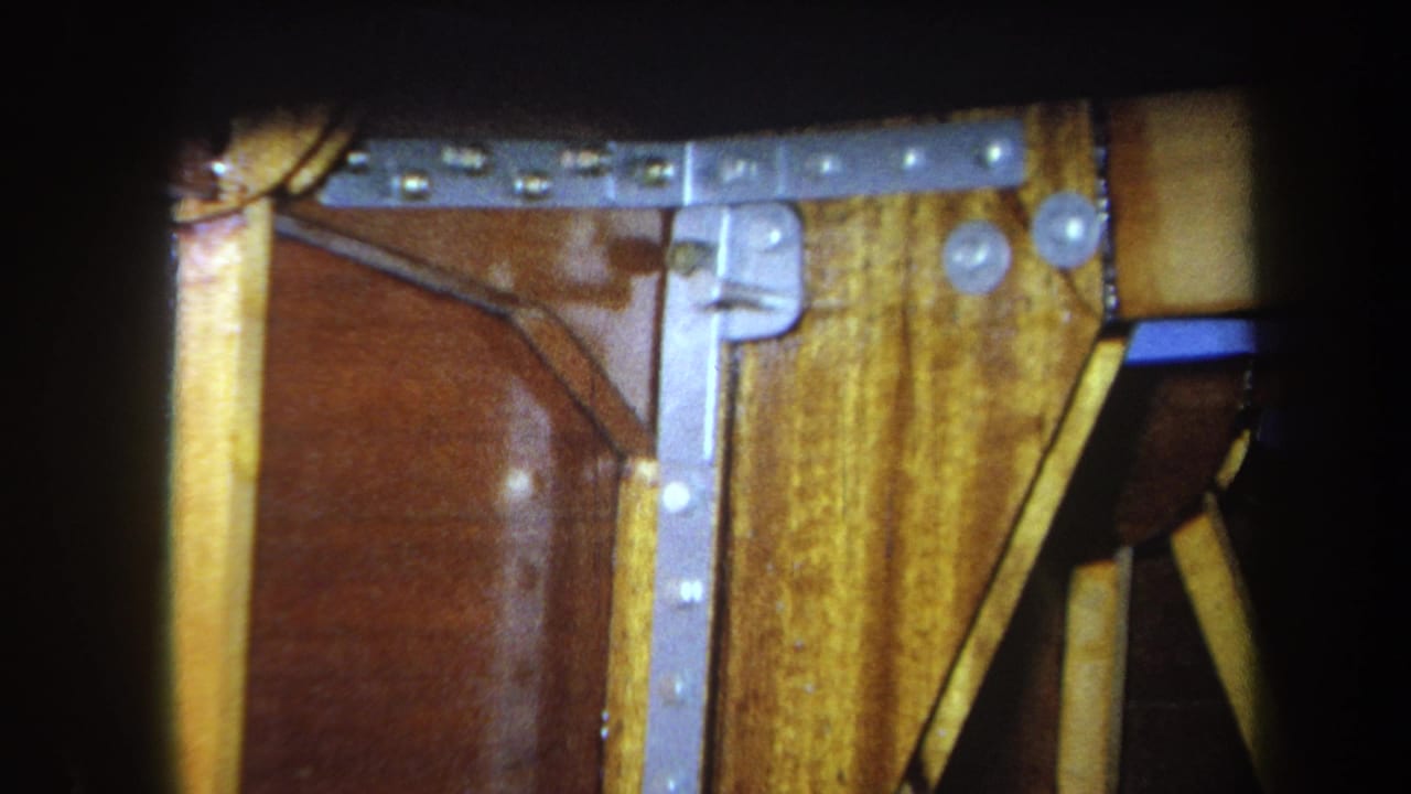 1964 Yucaipa 8mm frame showing a silver metal bracket, bolts, yellow boards, and brown varnished wood in close-up.