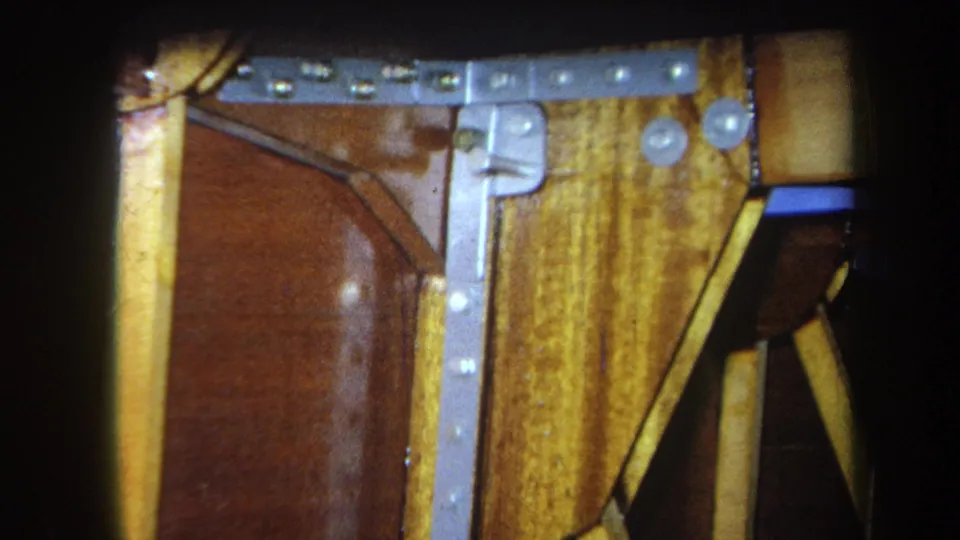 1964 8mm construction detail from Yucaipa with a bolted metal brace over yellow boards and a brown wooden surface.