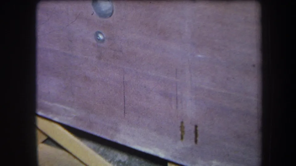 1964 home-movie frame showing a stained picnic table surface in soft focus, with water marks, scratches, and a pale board edge.