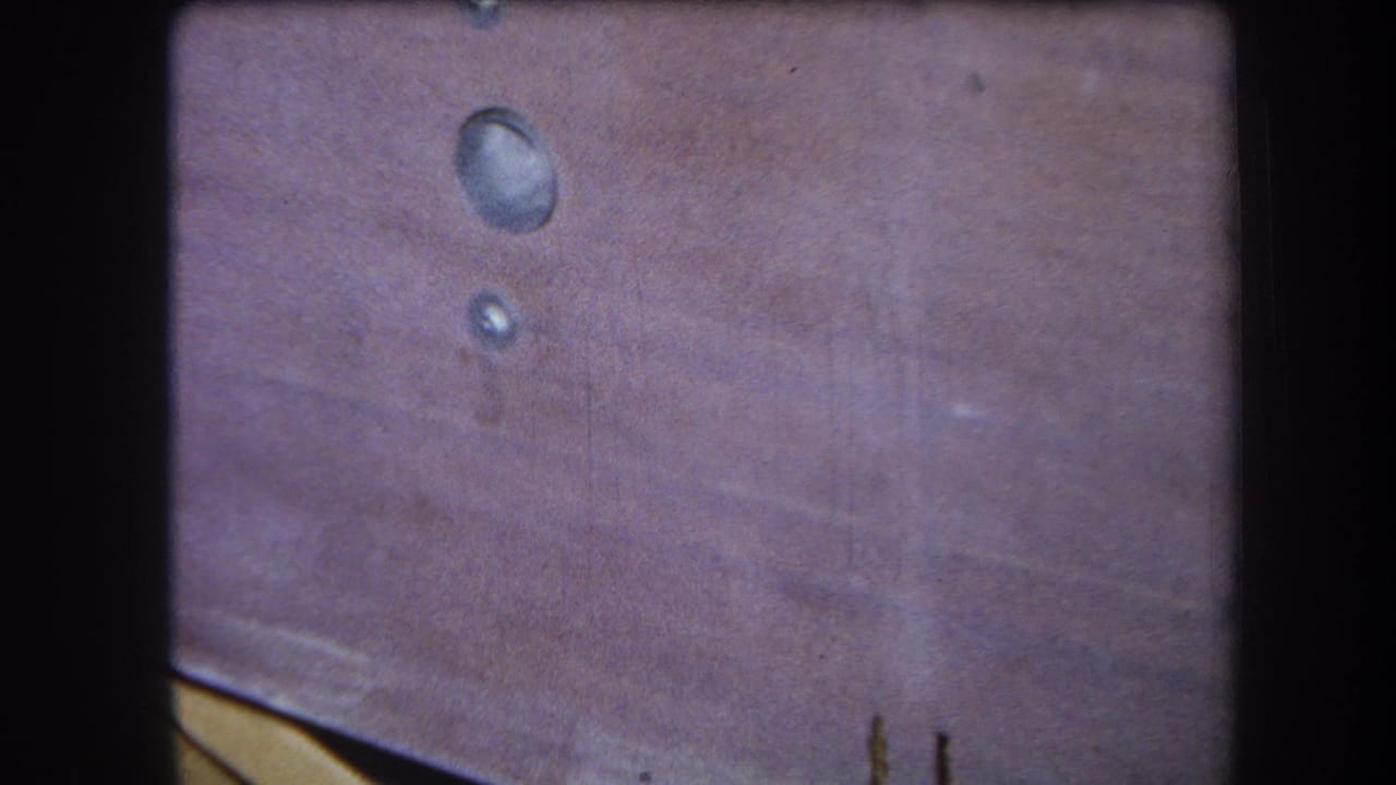 1964 8mm close-up from Yucaipa showing a pink-brown wooden surface with two reflective drops and visible film gate edges.