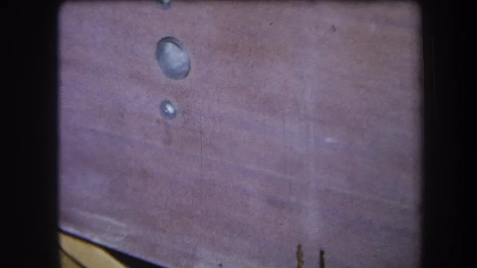 1964 8mm close-up from Yucaipa showing a pink-brown wooden surface with two reflective drops and visible film gate edges.